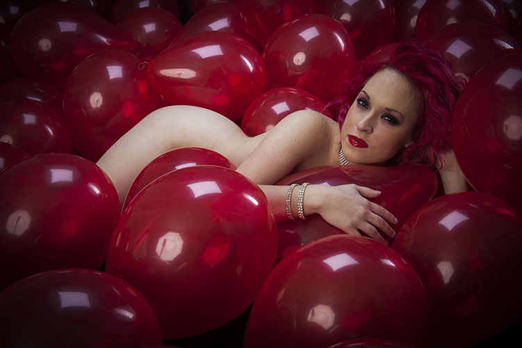 Implied nude female model laying in red balloons in photo studio