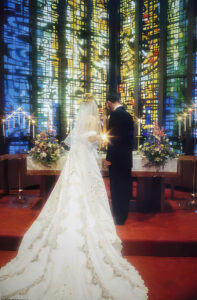 A bride and groom lighting a unity candle in front of large stained glass windows